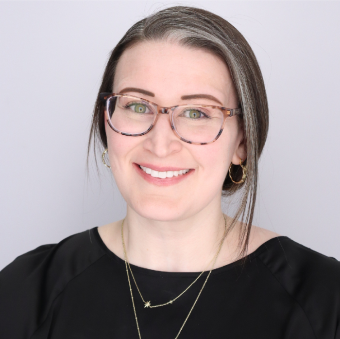A headshot photo of Dr. Lauren King. She is wearing a black shirt, two gold necklaces, large tortoise shell glasses, and has her hair back behind her head.